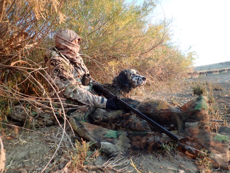 A hunter in camouflage sits with a dog in some bushes