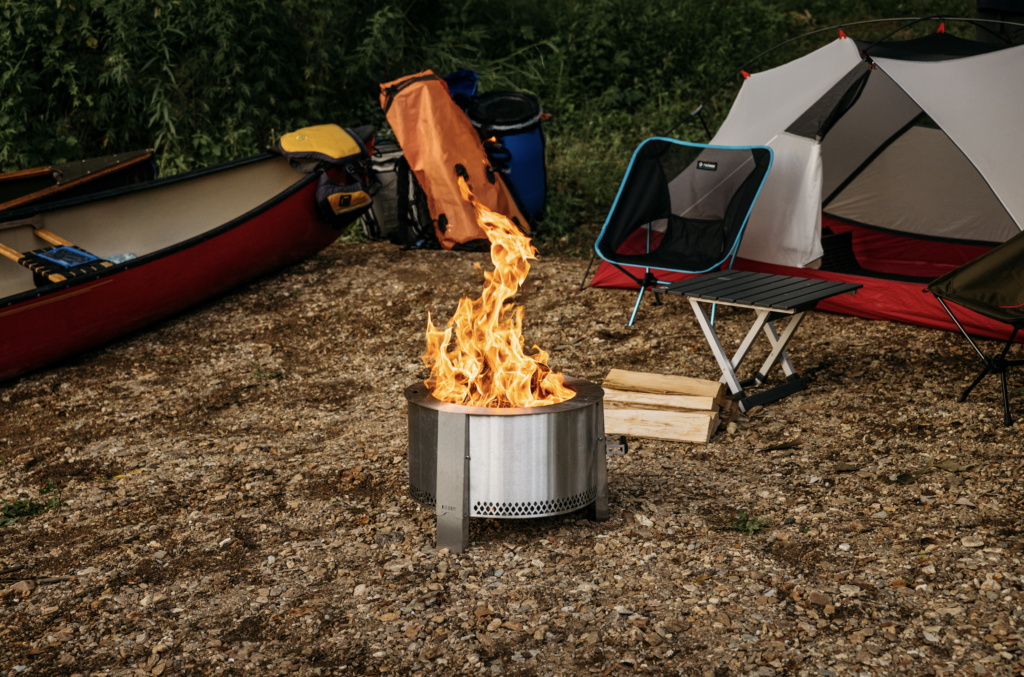 Breeo Y Series fire pit with fire set up at a base camp with a tent and chair and canoes.