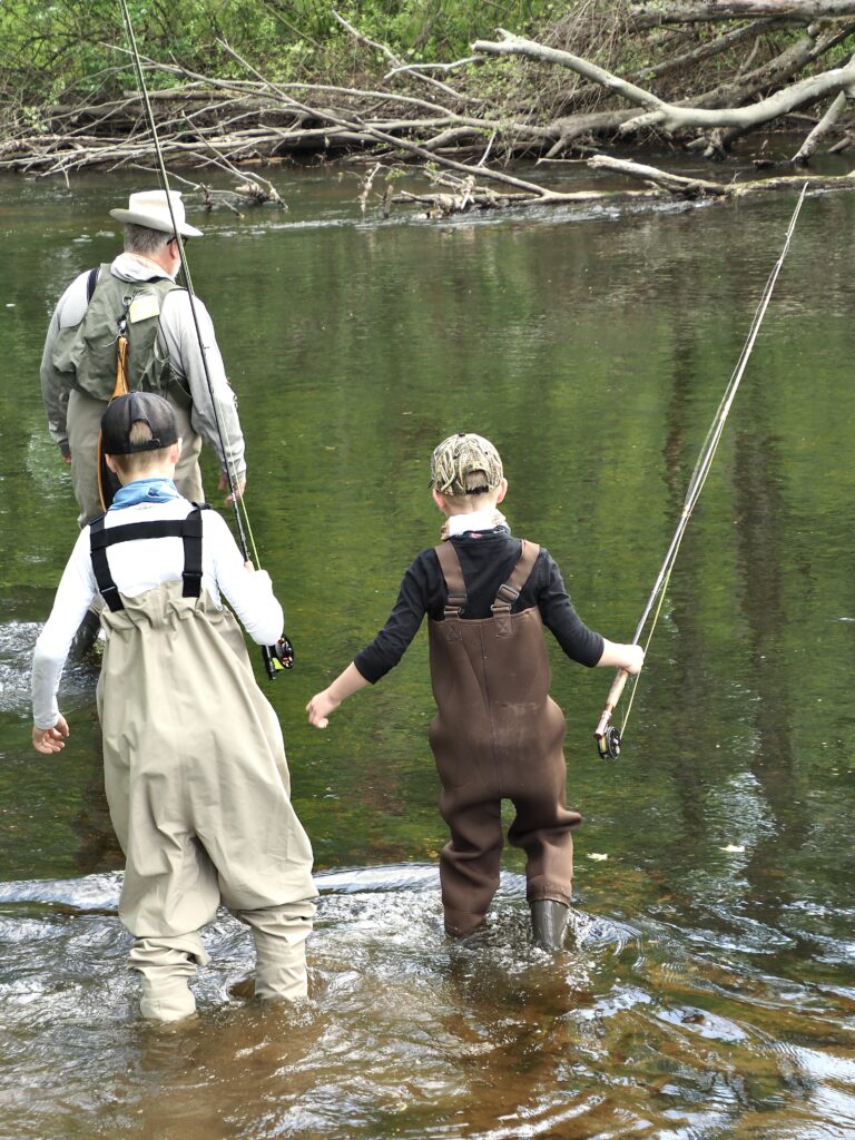An older man in hipwaders leads two children with fishing rods in the water
