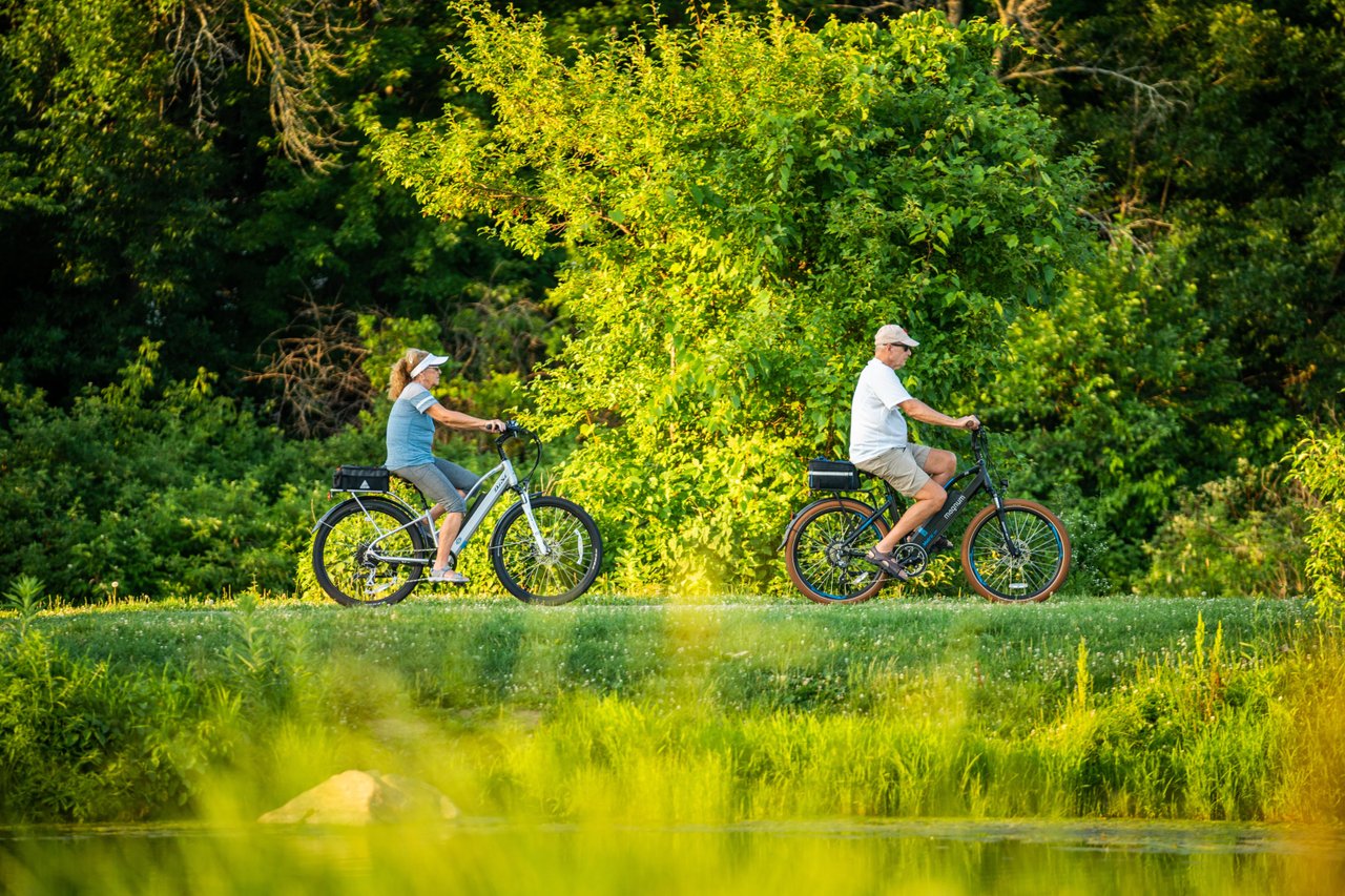 Two older people, seen in profile, bike along a flat trail surrounded by green