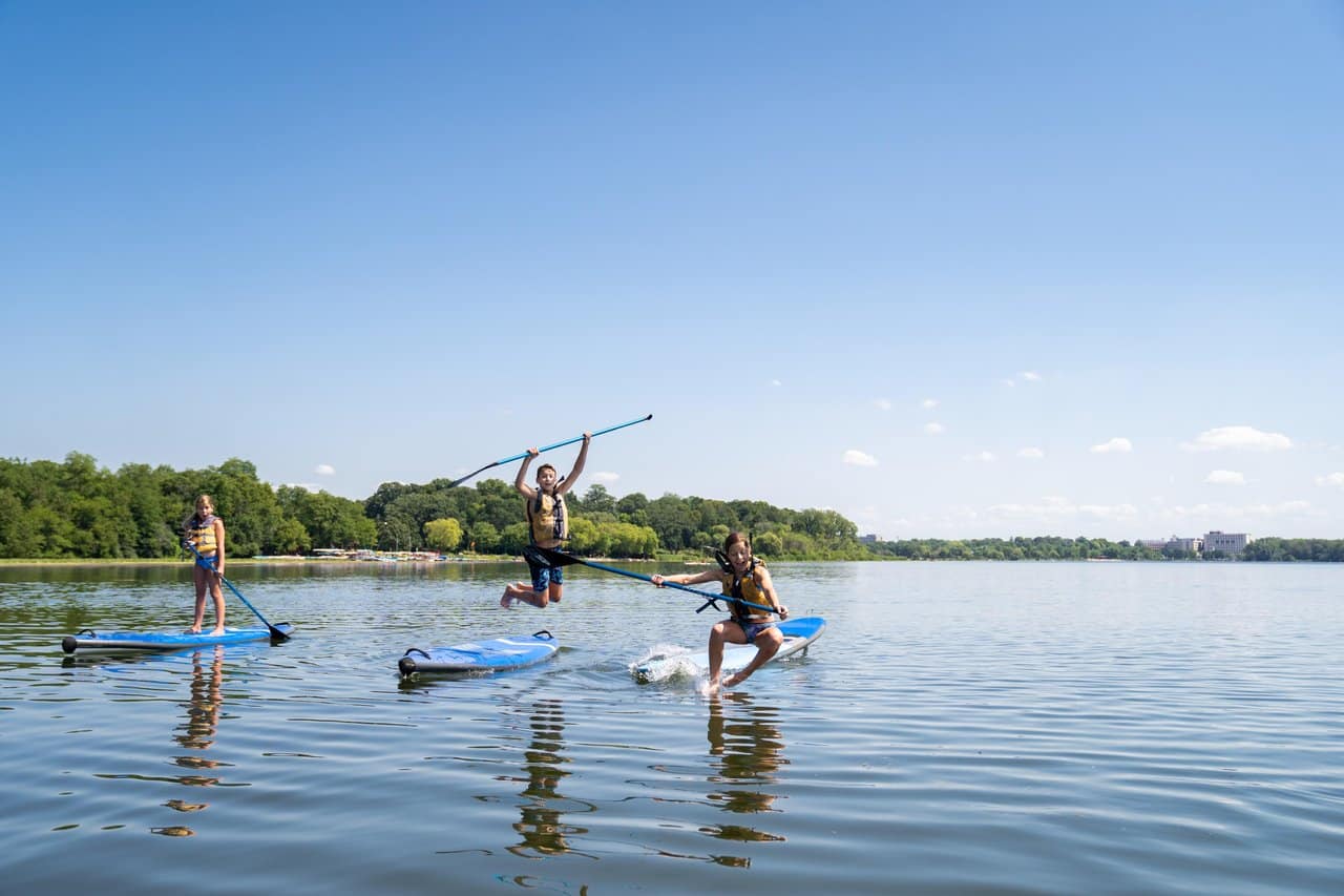 Three young people on paddleboards on a quiet lake; one of them leaps off the board with the paddle over their head