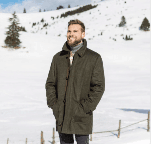 A man with a snowy background smiling wearing the longer style wool Loden Field Coat.