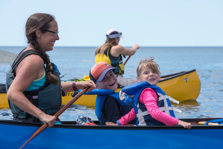 A person paddles a blue canoe with two children in front; a yellow canoe is seen in the background
