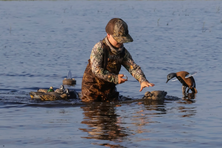 A child dressed in hunting gear stands in the water, reaching for a bird